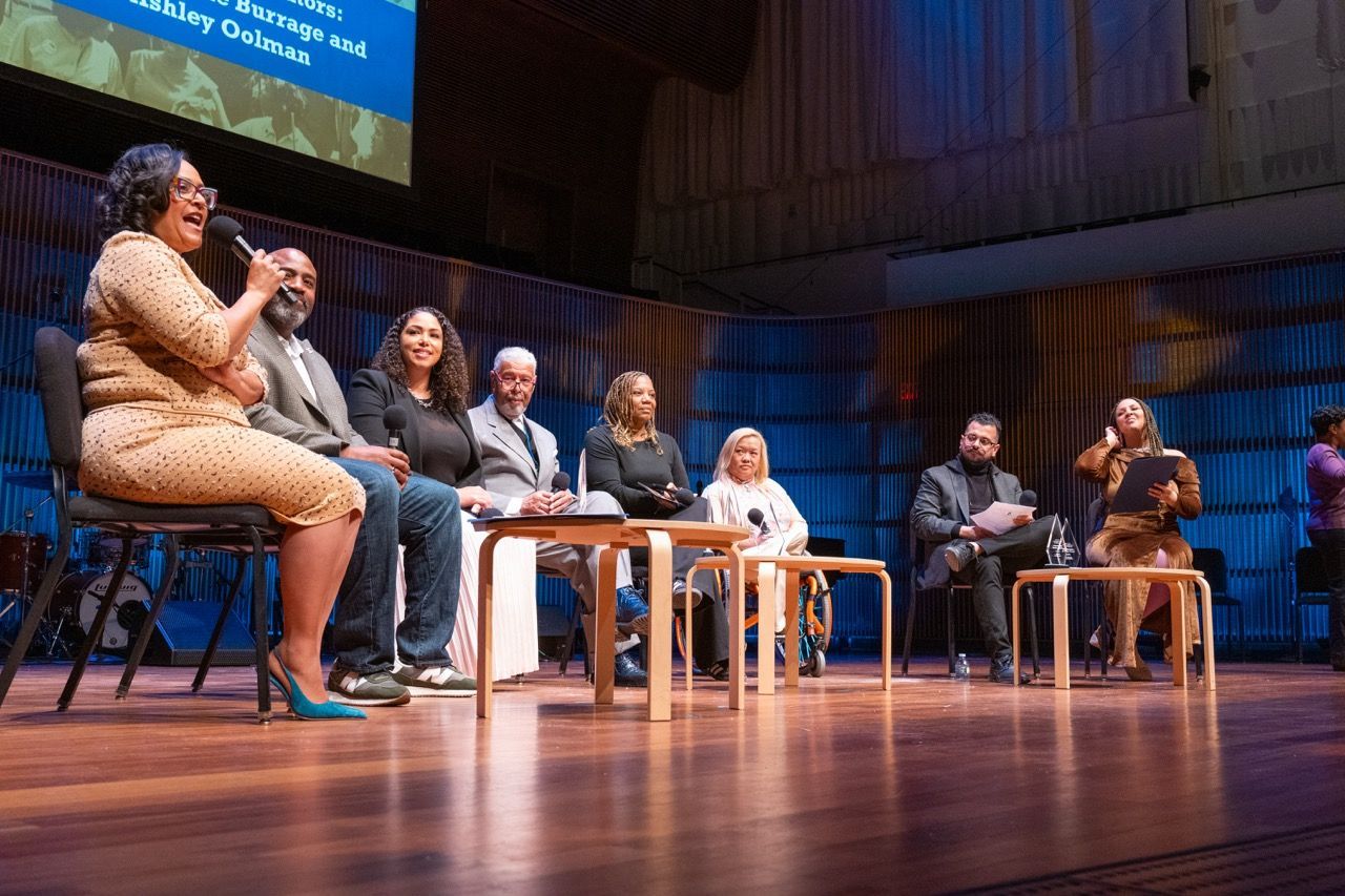 Panel discussion with eight diverse speakers seated on stage in a modern auditorium, speaking into microphones and engaging with the audience.